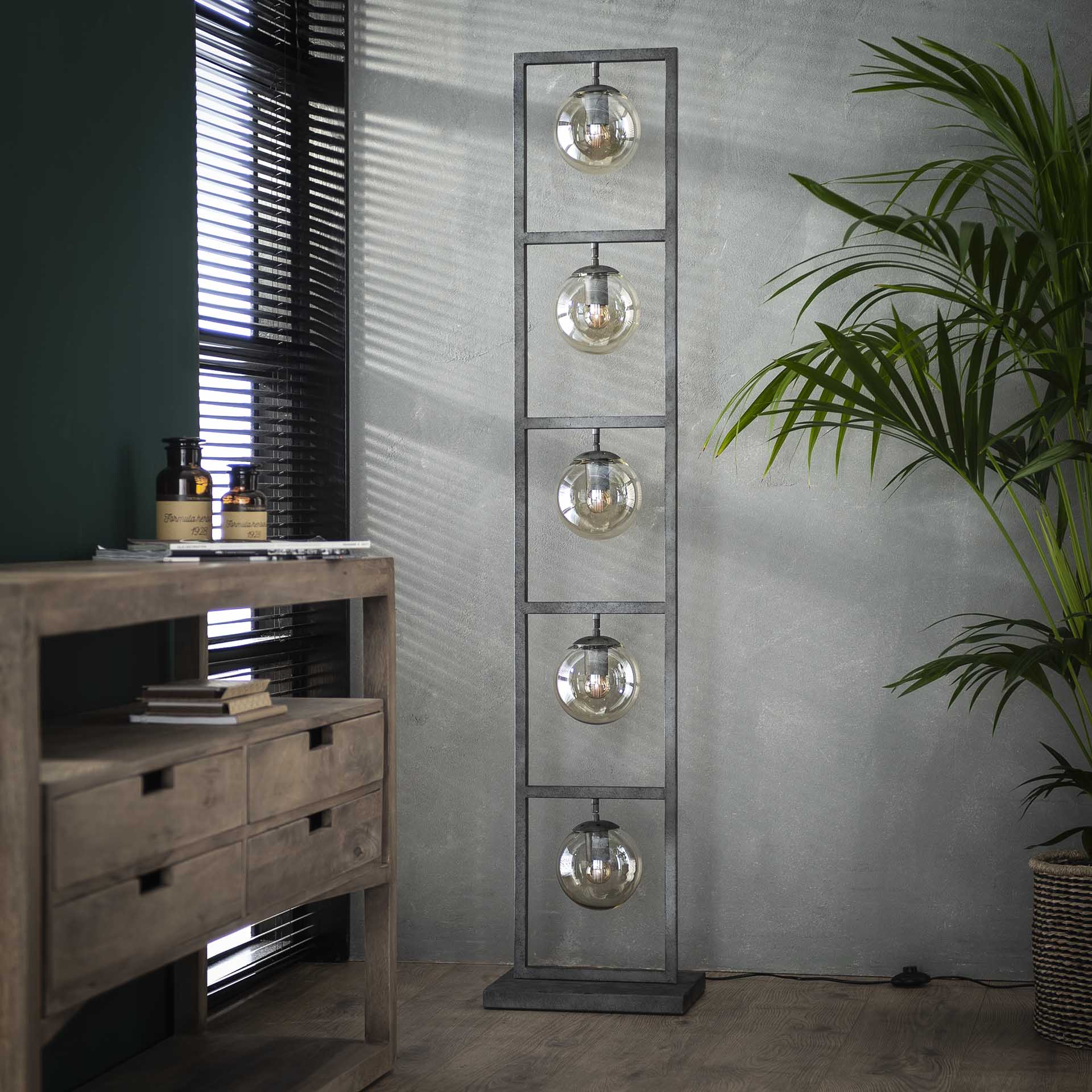 Industriele vloerlamp met metalen frame en glazen bollen naast houten meubel en plant in een woonkamer bij Belga Meubel Brugge