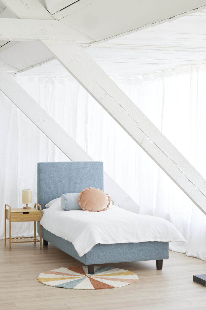 Slaapkamer met een blauw gestoffeerd bed, wit beddengoed, kleurige sierkussens, nachtkastje en sfeervolle houten balken