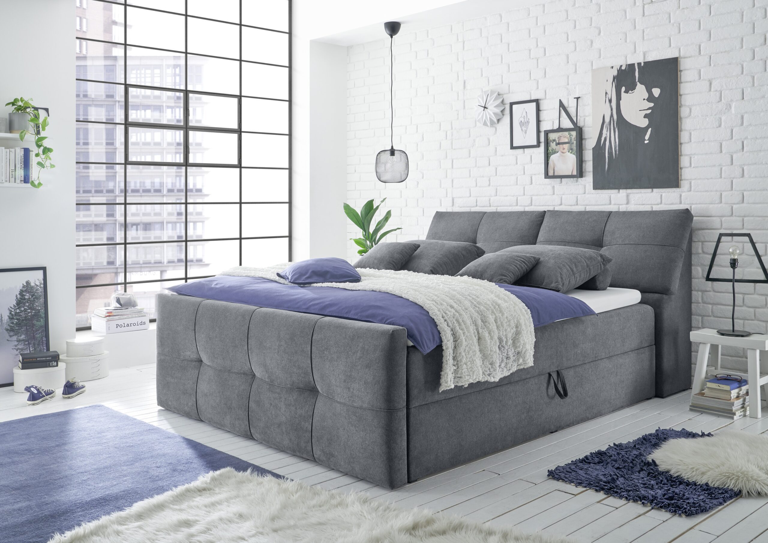 Slaapkamer met grijs boxspringbed, paars beddengoed, witte stenen muur en grote ramen bij Belga Meubel Brugge