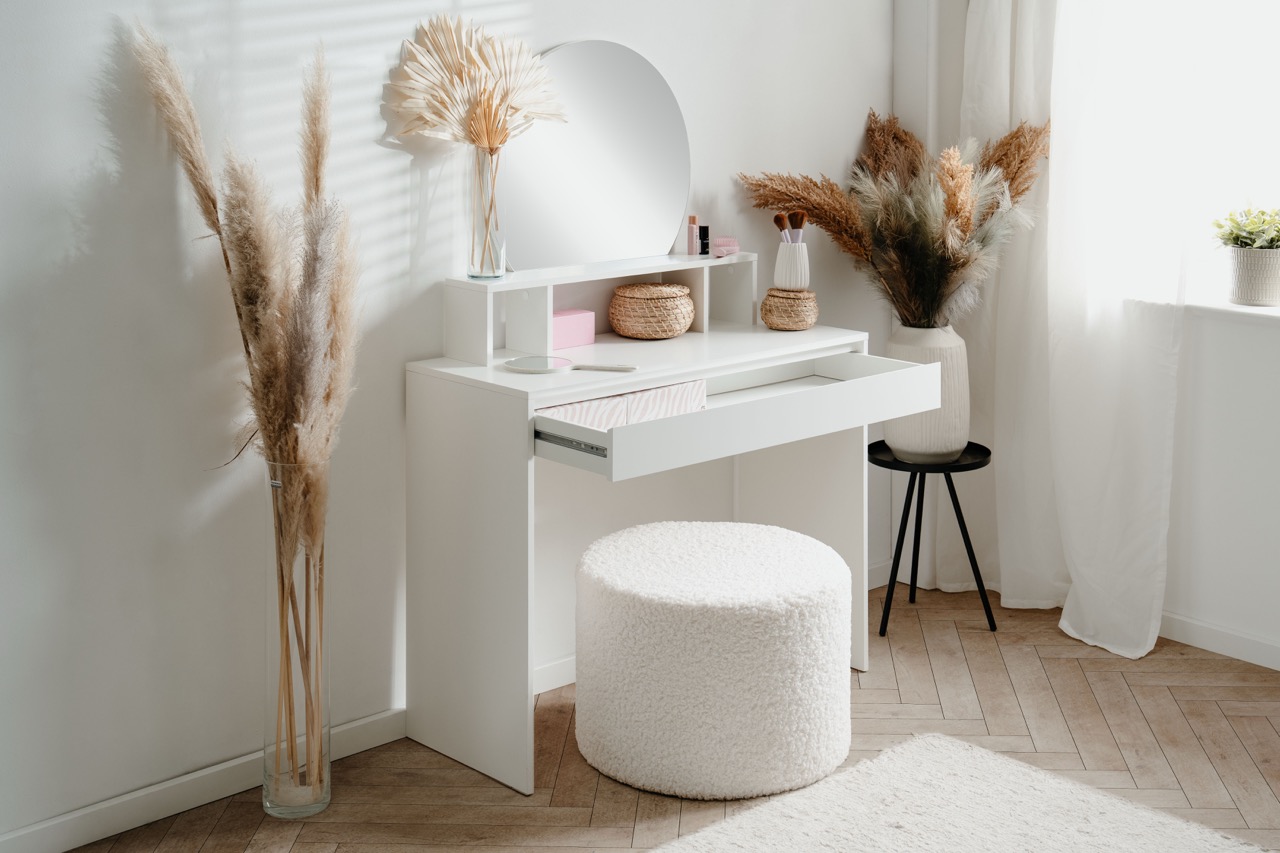 Witte make-up tafel met open lade, ronde spiegel en zacht poefje in lichte kamer met gedroogde bloemen en houten vloer